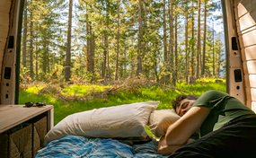 Ein Mann schläft auf einem Bett in einem Van, dessen geöffnete Hecktüren einen Blick auf einen sonnigen Wald freigeben.