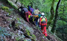 Plusieurs personnes transportent une personne sur une civière orange à travers une pente raide et boisée. Les secouristes portent des vêtements d'intervention rouges et jaunes et des casques.
