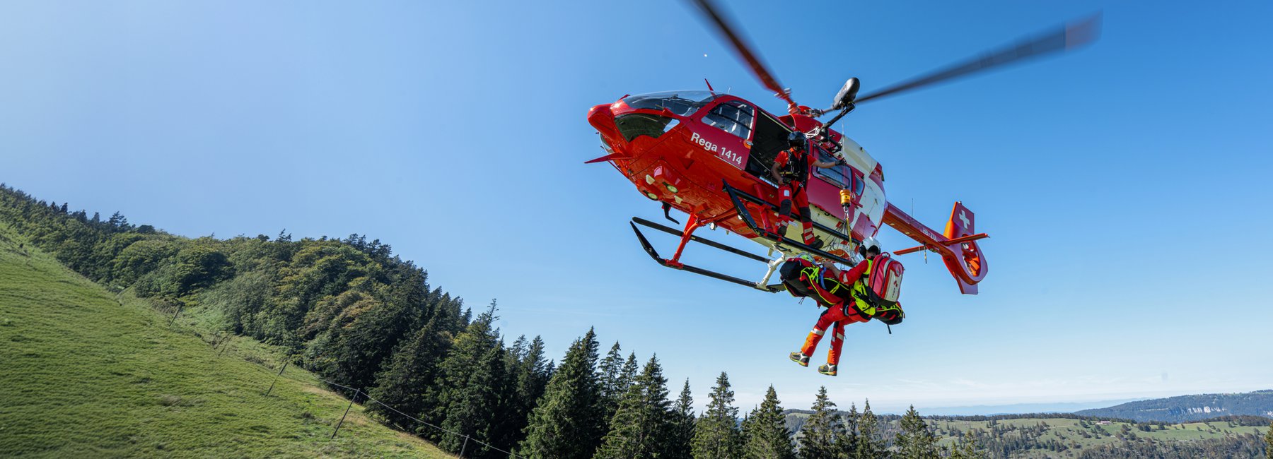 Ein Rega-Helikopter im Schwebeflug. Ein Rettungssanitäter wird mit einem Patienten am Haken unter dem Helikopter abgeseilt. Im Hintergrund ist eine bewaldete Landschaft zu sehen.