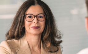 Eine professionell gekleidete Frau, die Brille trägt und einen beigefarbenen Blazer anhat, sitzt an einem Tisch. Sie blickt direkt in die Kamera, während ein junger Mann mit einem weissen Kapuzenpullover im Vordergrund mit dem Rücken zur Kamera sitzt. Auf dem Tisch steht ein offener Laptop.