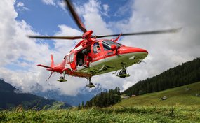Un hélicoptère de sauvetage AgustaWestland Da Vinci de la Rega est en vol stationnaire à basse altitude au-dessus d'une prairie de montagne. Un spécialiste en sauvetage de la Rega se tient dans la porte latérale ouverte et regarde vers le bas. Un paysage de montagne boisé est visible en arrière-plan.