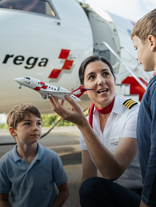 Eine Rega-Jet-Pilotin in Uniform kniet vor zwei Kindern und zeigt ihnen ein Modellflugzeug des Rega Challenger 650 Ambulanzjets. Im Hintergrund ist die offene Tür eines echten Rega Ambulanzjets sichtbar.