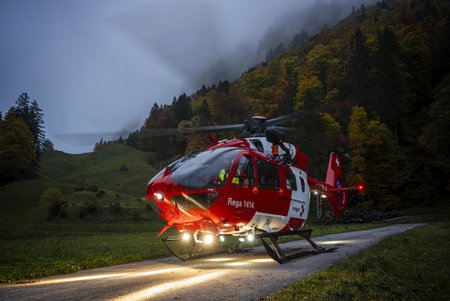 Zum Downloadformular für das Bild Rega-Helikopter mit eingeschaltetem Suchscheinwerfer bei einem Einsatz in hügeligem Gelände.