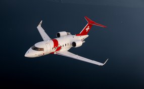 Un jet di soccorso Rega Challenger 650, con la sigla HB-JWA, vola a bassa quota sopra un lago calmo.