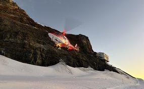 Ein Rega-Rettungshelikopter Da Vinci fliegt im frühen Morgengrauen über eine schneebedeckte Berglandschaft. Im Bild sind die beleuchteten Rotorblätter und das typische Rot-Weiss des Helikopters sichtbar. Im Hintergrund ist eine Berghütte zu sehen.