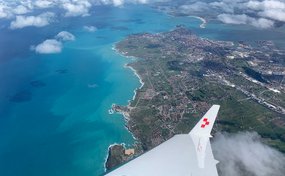 Vista da un jet ambulanza della Rega su una costa con mare turchese, campi verdi e una città sotto uno strato di nuvole.