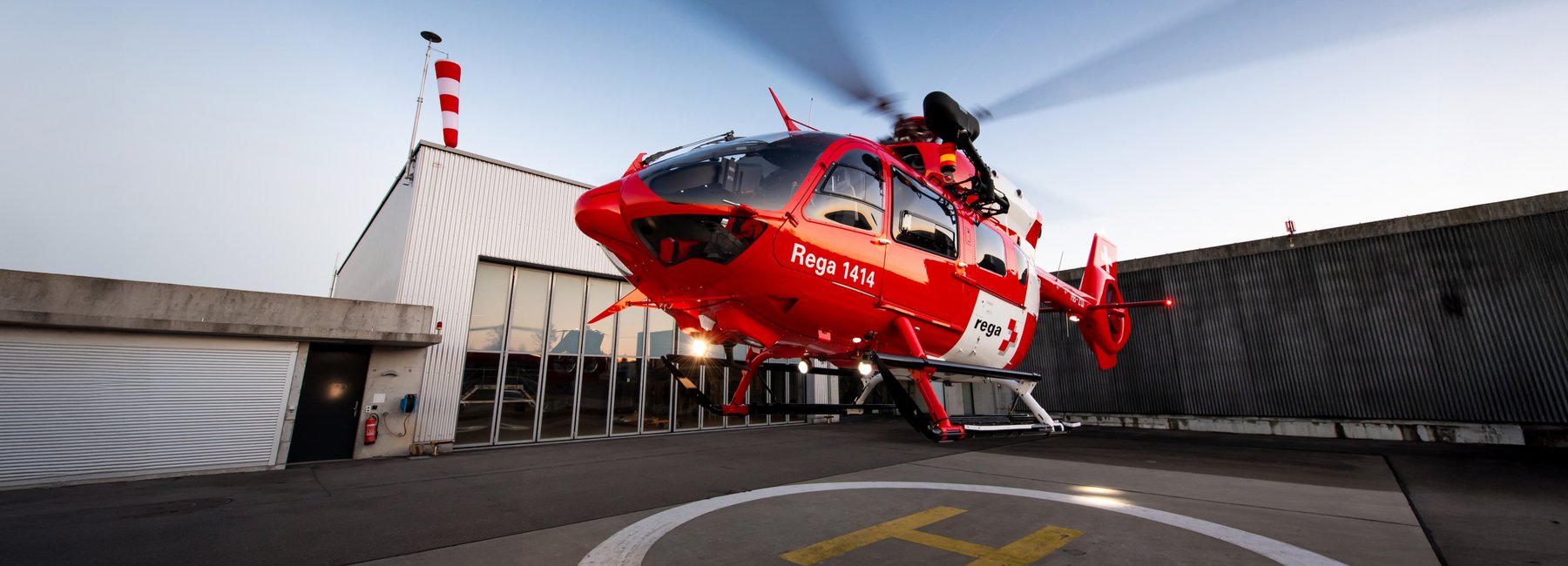 Ein Rega-Helikopter hebt von der Basis ab. Im Hintergrund ist ein Hangar zu sehen.