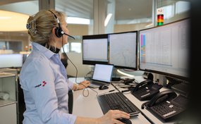 Une médecin-conseil de la Rega portant une chemise blanche et bleue est assise au centre d'opérations devant trois moniteurs d'ordinateur. Elle porte un casque et utilise une souris pour gérer les informations sur les écrans et répondre aux appels d'urgence.