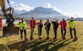 Group photo at the groundbreaking ceremony for the new Rega base in Samedan. Seven individuals, some in red Rega operational gear and some in suits or workwear, stand side-by-side with spades on a meadow in front of construction machinery and a snow-capped mountain backdrop.