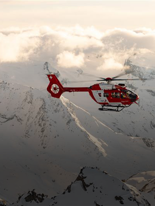 Ein Rega Airbus H145 Rettungshelikopter fliegt in geringer Höhe über eine majestätische, schneebedeckte Berglandschaft. Der rot-weisse Helikopter taucht zwischen felsigen Gipfeln und Wolken auf.