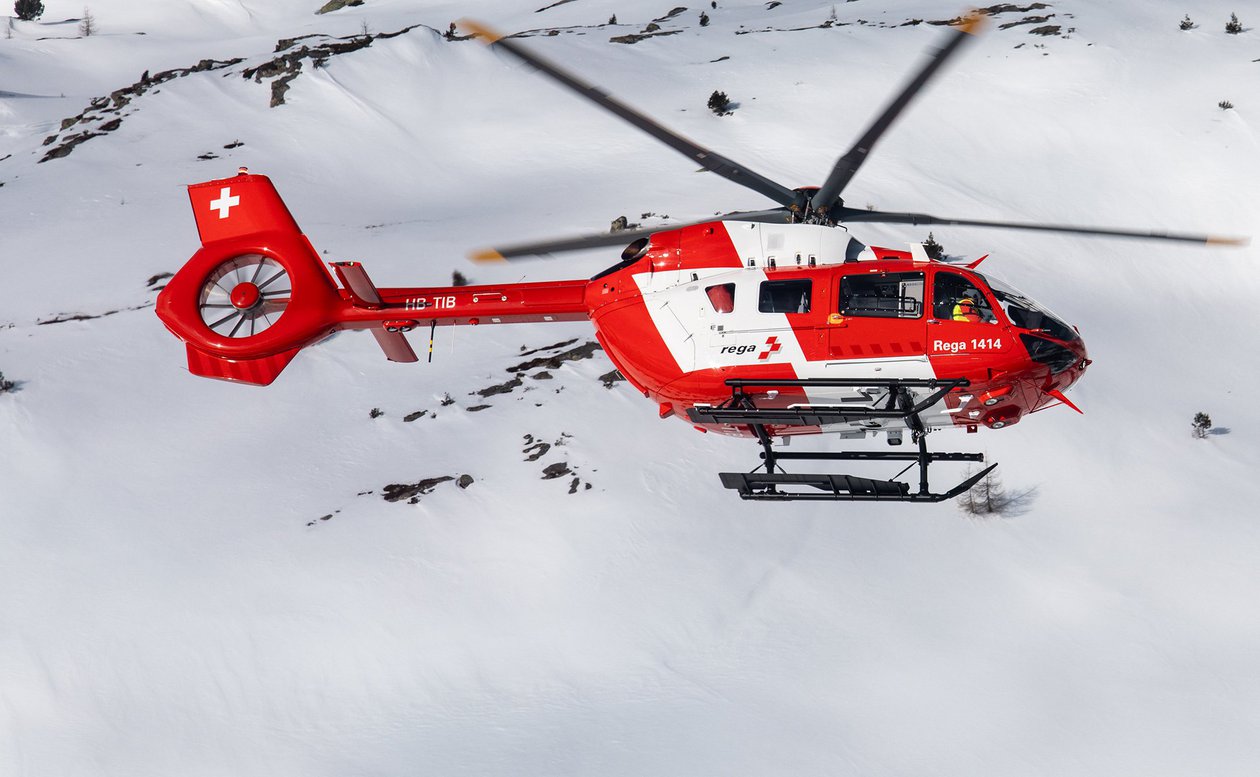 Ein roter und weisser Rega Airbus H145 Helikopter mit der Immatrikulation HB-TIB fliegt über eine weitläufige, tief verschneite und unbewaldete Berglandschaft. Das Bild zeigt den Helikopter bei einem Einsatz im Winter.