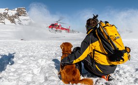 Lawinenhundeführer und sein Lawinenhund warten im Schnee bis der Rega-Helikopter gelandet ist