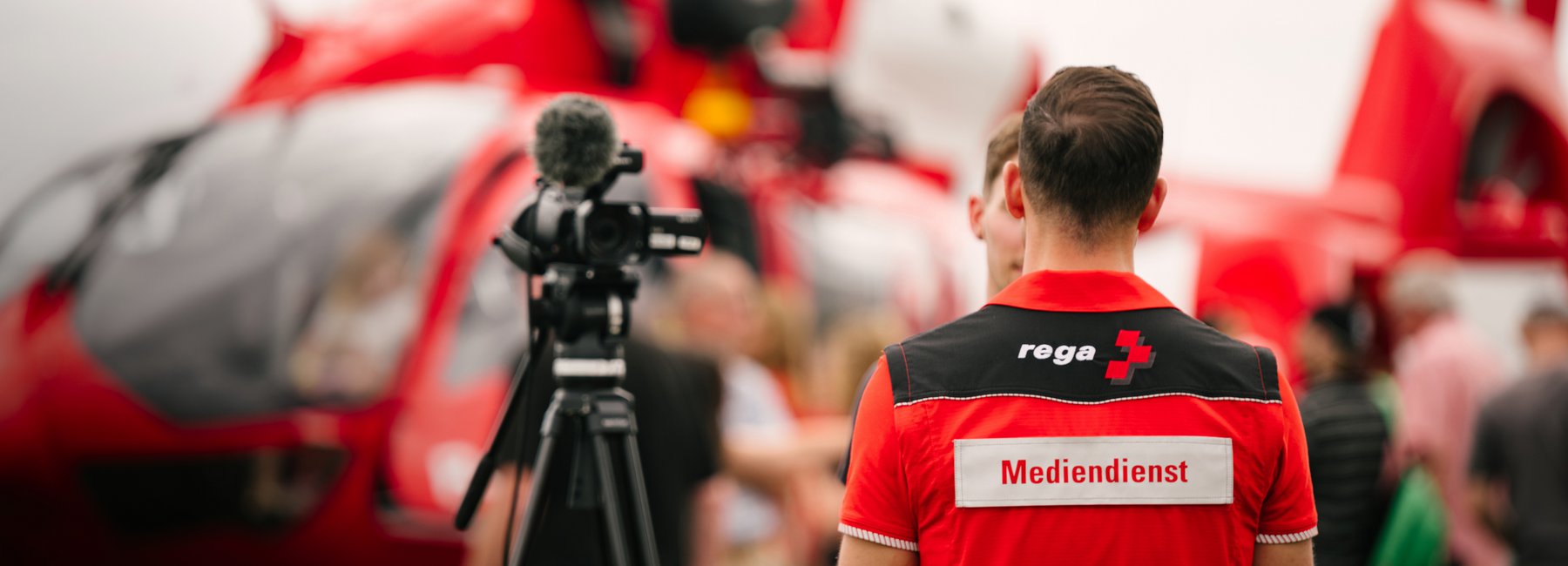 Ein Rega-Mitarbeitender steht mit dem Rücken zur Kamera vor einem Helikopter. Auf seinem Rücken steht «Mediendienst». Neben ihm ist eine Videokamera auf einem Stativ.