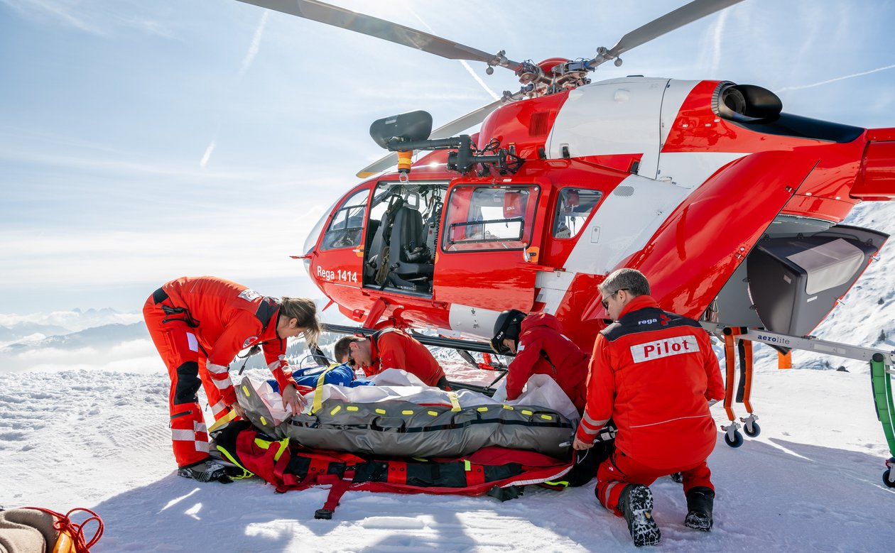 Eine Rega-Crew (Pilot und medizinische Crew) versorgt einen Patienten auf einer Schneeunterlage neben einem roten und weissen Airbus H145 Helikopter in einer weissen, sonnigen Berglandschaft.