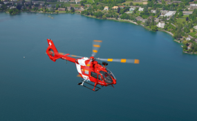 Ein roter Rega-Helikopter fliegt über einem See mit bewaldeten Ufern und Siedlungen im Hintergrund.
