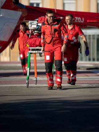 Zwei Rega-Mitarbeitende in roter Uniform transportieren eine Trage mit medizinischer Ausrüstung, während ein Helikopter im Hintergrund steht.