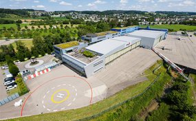 Eine Luftaufnahme des Rega-Centers am Flughafen Zürich-Kloten. Der moderne, mehrteilige Gebäudekomplex mit weissen Hangars und begrüntem Dach ist umgeben von einer Landebahn mit einem deutlich sichtbaren Helikopterlandeplatz. Im Hintergrund ist eine grüne Landschaft mit einem Dorf zu sehen.
