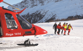 Un equipaggio Rega trasporta una persona su una barella attraverso un paesaggio montano innevato verso l'elicottero di soccorso pronto al decollo.