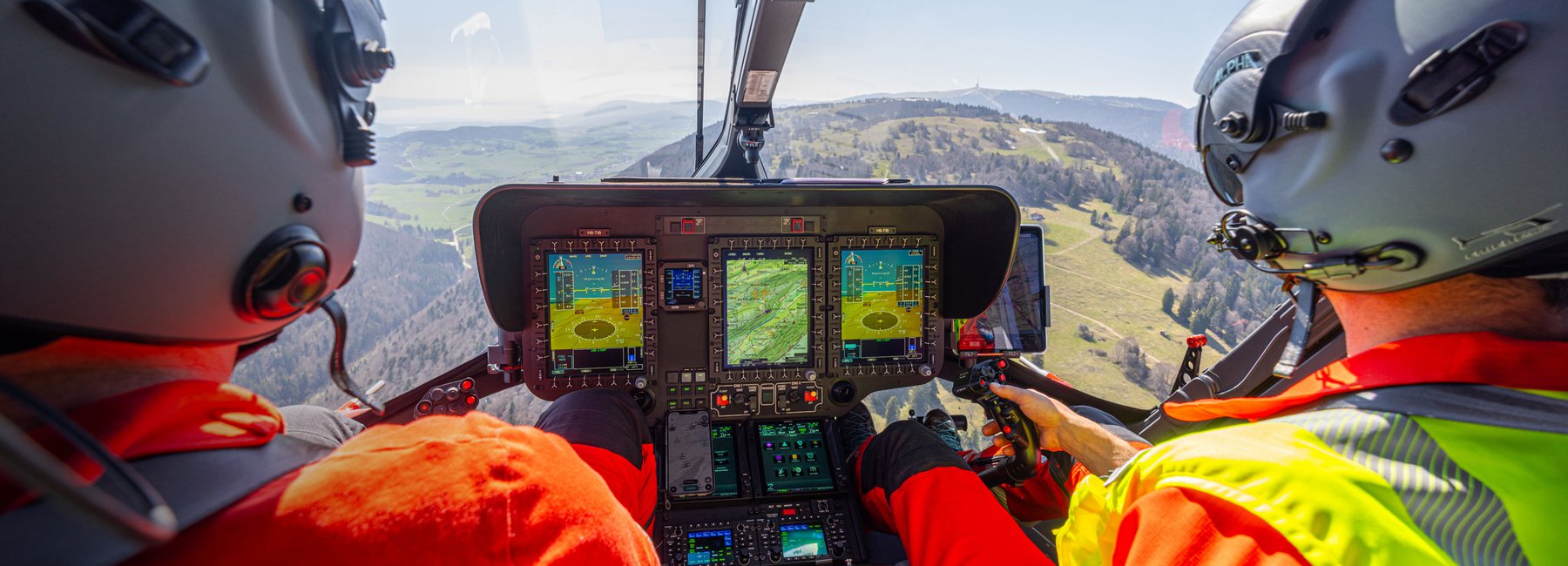 Ansicht aus dem Cockpit eines Rega-Helikopters während des Fluges, mit zwei Piloten und dem Bedienfeld. Im Hintergrund ist eine bewaldete Hügellandschaft zu sehen.