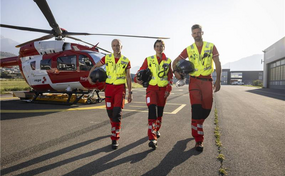 Drei Rega-Crewmitglieder gehen in ihren roten Einsatzanzügen und gelben Warnwesten auf dem Helikopterlandeplatz. Im Hintergrund steht ein Rega-Helikopter vom Typ Airbus H145.