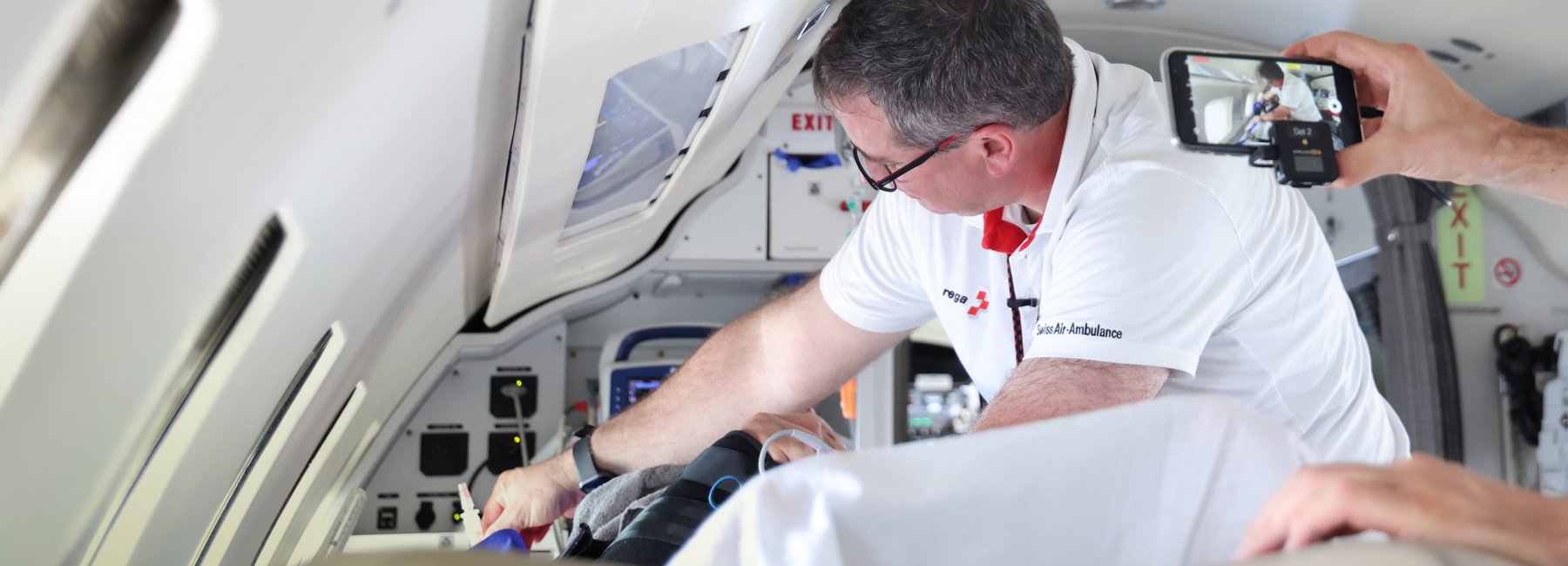 Eine Person in Rega-Kleidung wird beim Arbeiten in einem Rega-Ambulanzjet von einem Smartphone gefilmt.