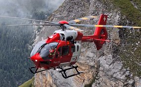 Un hélicoptère de sauvetage de la Rega vole près d'une paroi rocheuse abrupte dans les montagnes. Il a la livrée rouge et blanche typique de la Rega. Les membres de l'équipage sont visibles sur le côté gauche de l'image.
