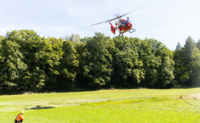 Un hélicoptère Rega H145 vole à basse altitude au-dessus d'une prairie. Un secouriste agenouillé dans l'herbe regarde l'hélicoptère. Un groupe d'arbres est visible en arrière-plan.