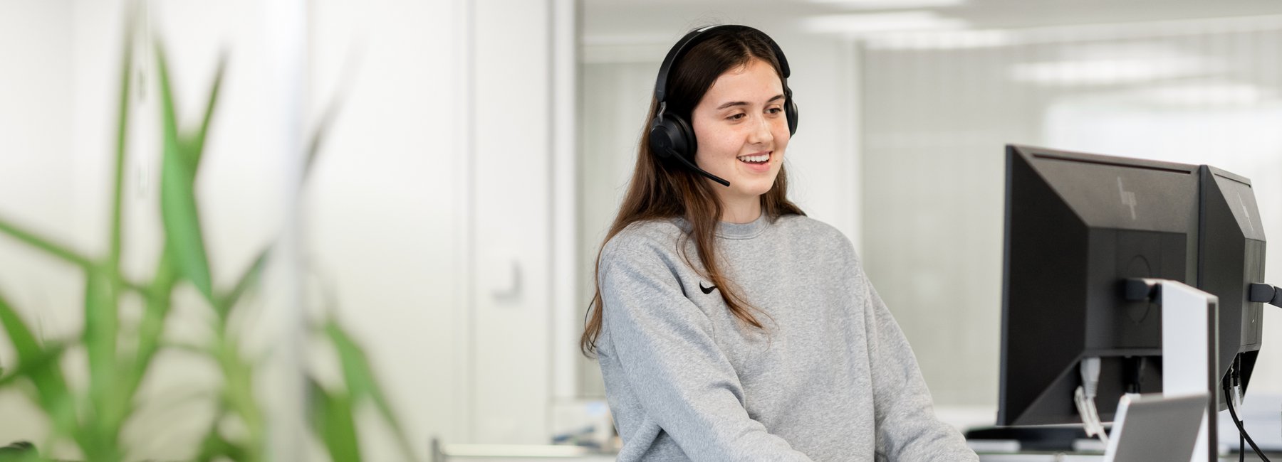 Eine lächelnde junge Frau mit Headset sitzt an einem Schreibtisch und arbeitet an einem Computer. Sie repräsentiert die Einsatzleiterinnen und -leiter in der Einsatzzentrale.
