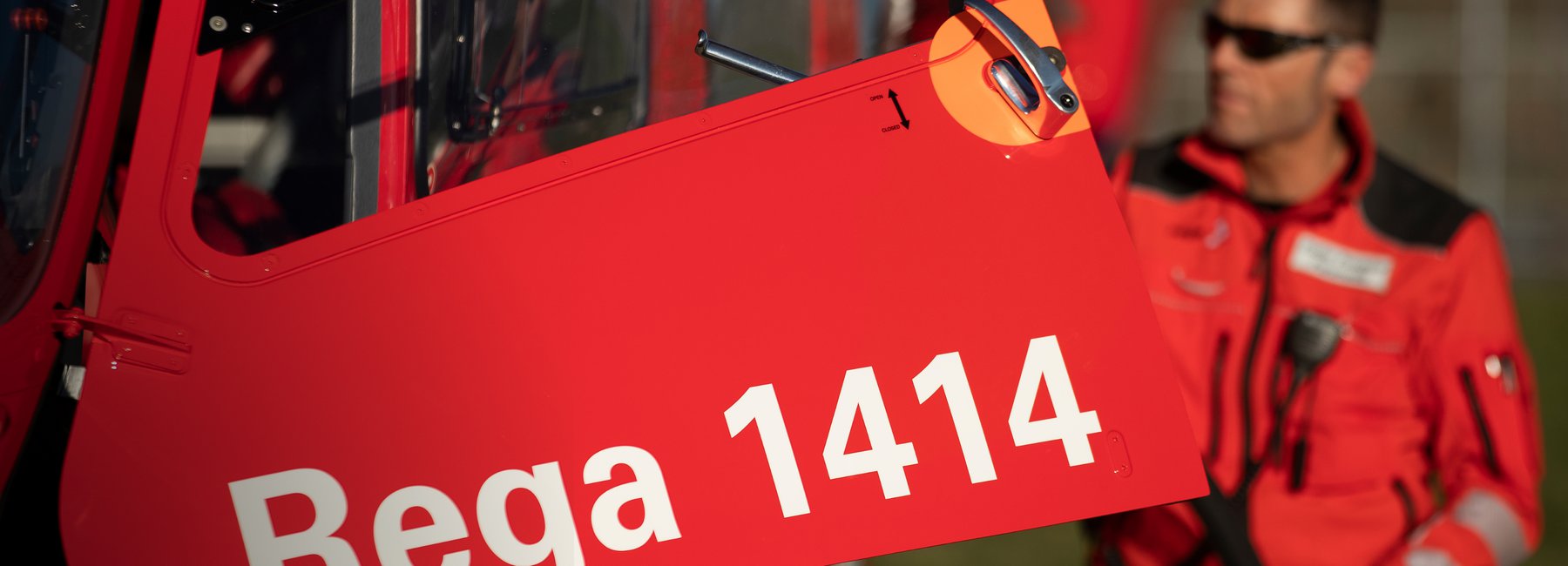 Nahaufnahme eines roten Rega-Helikopters. Auf der Seitentür steht die Notrufnummer 1414. Im Hintergrund ist eine Person in roter Uniform unscharf zu erkennen.