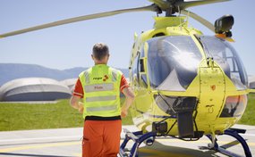 A Rega pilot, wearing an orange uniform and a yellow high-visibility vest with the inscription "rega Pilot," stands with his back to the camera, approaching a yellow rescue helicopter on a landing pad.